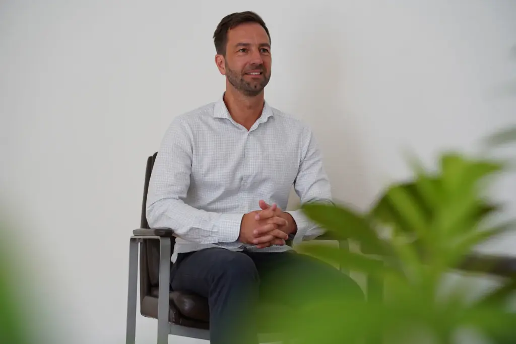 life-coaching-hero-thomas-waaden Thomas Waaden sitzt ruhig in einem hellen Raum, im Vordergrund eine grüne Pflanze.